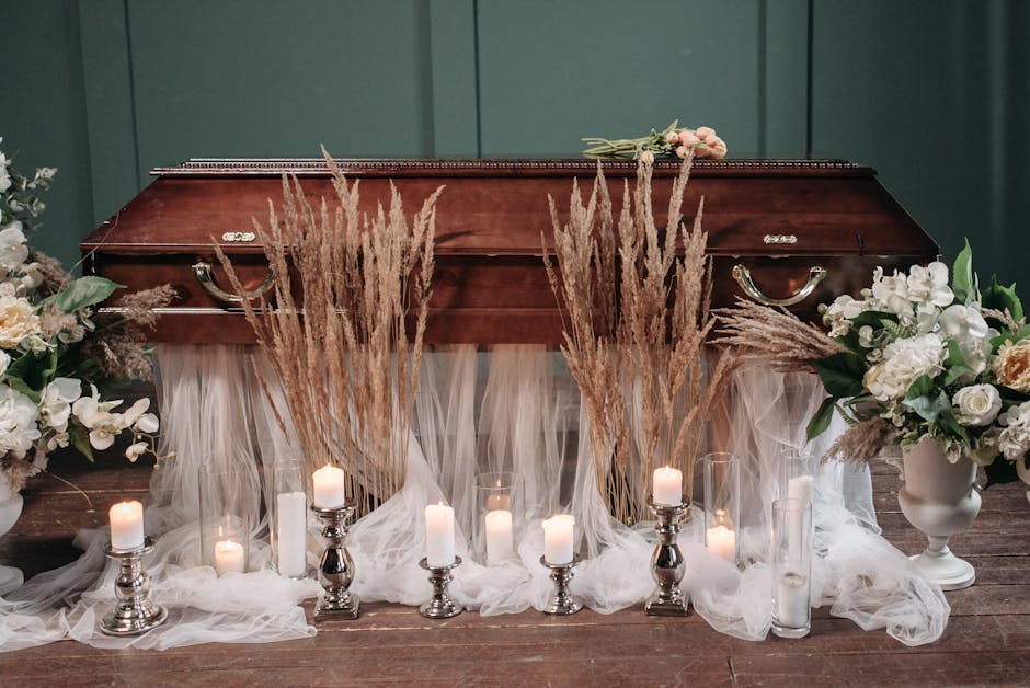 A wooden funeral casket decorated with an arrangement of predominantly white and cream flowers, including roses and lilies, flanked by green foliage. Surrounding the casket are tall, dried pampas gras