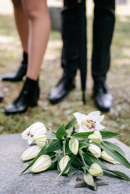 A bouquet of white lilies with large, open petals and prominent stamens, surrounded by smaller lily buds and elongated green leaves, resting on a stone surface. In the background, the lower legs and f