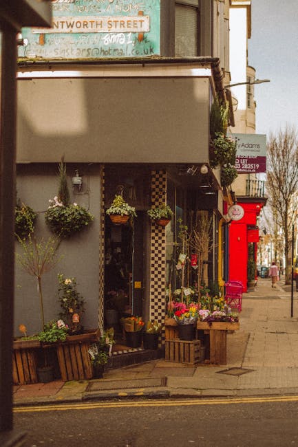 A street-level view of a florist shop featuring a variety of freshly cut flowers arranged in baskets and pots outside the entrance, including colorful blooms such as pink, purple, and yellow flowers.