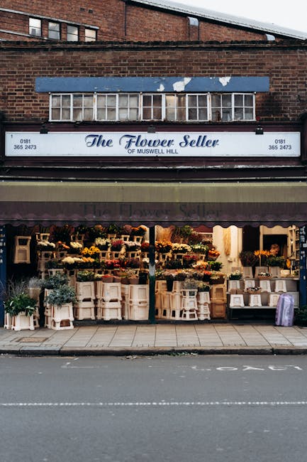 The storefront of The Flower Seller of Muswell Hill features a display of fresh floral arrangements arranged on multiple white shelving units and pedestals in a variety of vibrant colors, including ye