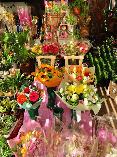 A display of fresh flower bouquets arranged on a pink cloth-covered table inside a florist shop. The bouquets feature a variety of flowers including bright pink gerbera daisies, yellow sunflowers, whi