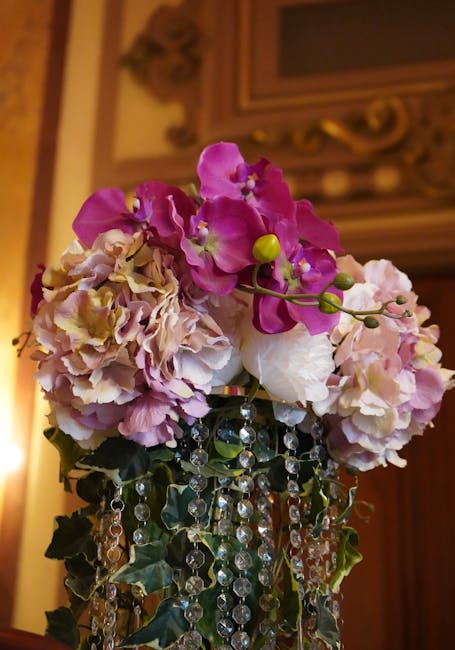 A floral arrangement featuring vibrant pink and cream hydrangeas with some green buds, complemented by purple orchids on long, curved stems. The bouquet is adorned with dangling strands of clear cryst