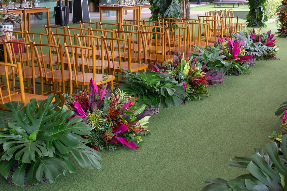 A floral arrangement featuring vibrant pink and purple flowers, including bromeliads and lilies, integrated with lush green foliage such as monstera leaves and ornamental grasses. The flowers are set